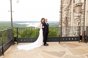 A bride and groom share an intimate moment on a scenic stone balcony overlooking rolling hills and mountains.