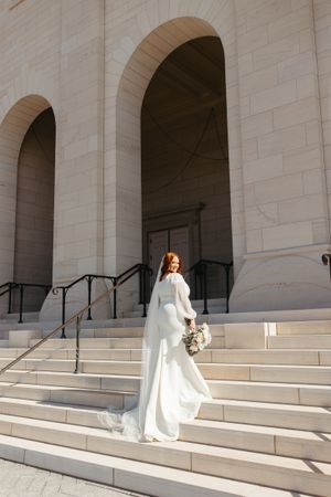 A bride in a flowing white dress poses elegantly on stone steps of a grand building.