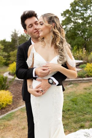 A romantic wedding couple embraces outdoors in a garden setting with greenery in the background.