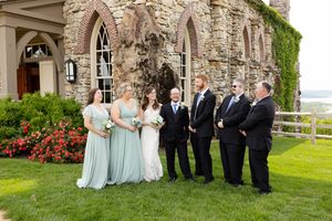 Wedding party poses outside stone castle venue with Gothic arched windows and flowering garden on sunny day.