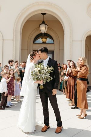 A wedding couple shares a romantic moment surrounded by guests under an ornate arched entryway.