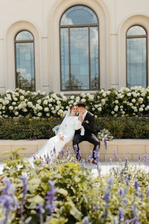 A couple shares an intimate moment near a white building with arched windows and beautiful white floral landscaping.