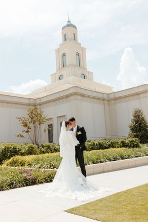 A couple poses for a wedding photo outside a beautiful white temple with a tower and manicured gardens.