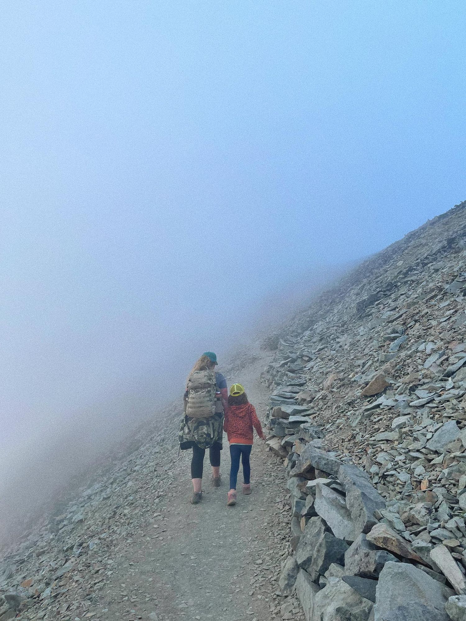 Two hikers walk along a misty mountain trail with rocky slopes and foggy conditions in the background.