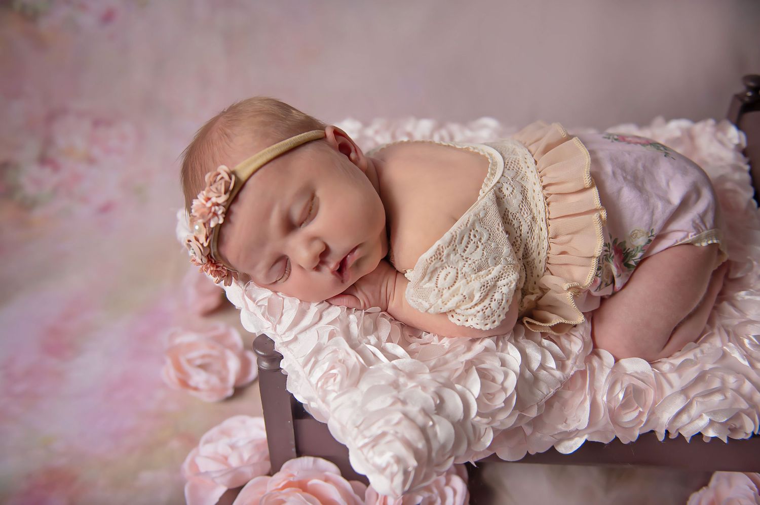 Sleeping newborn wearing a delicate lace outfit and floral headband on textured pink fabric.