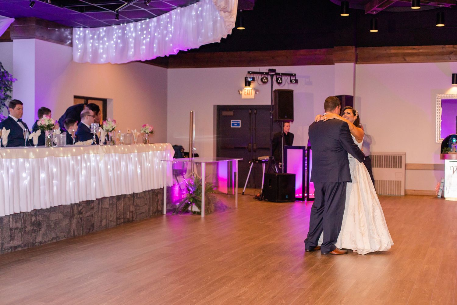 A couple dances in an elegantly lit reception hall with white draped head table and purple accent lighting.