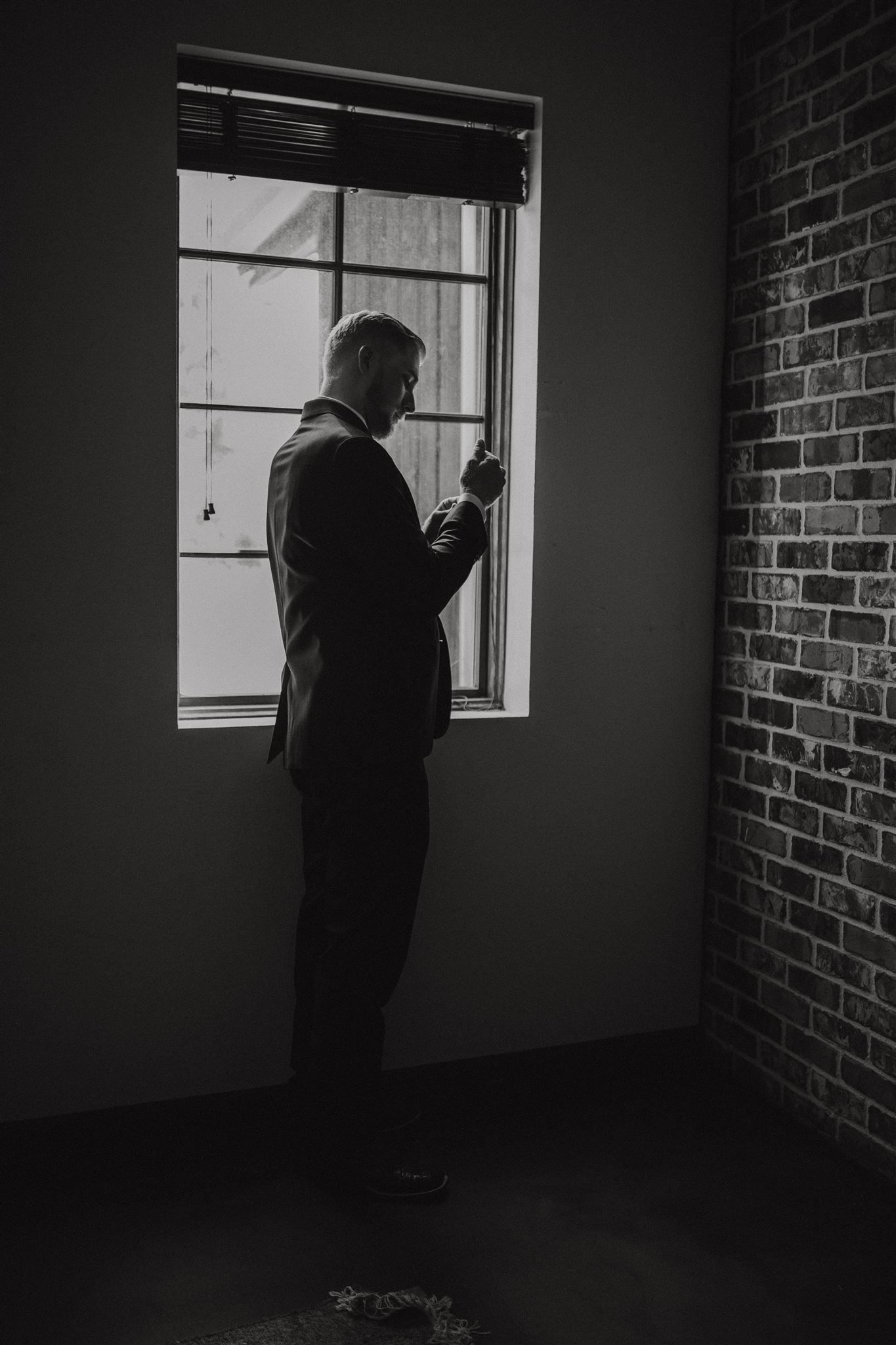 Silhouetted figure in formal attire stands by window against exposed brick wall in moody lighting.