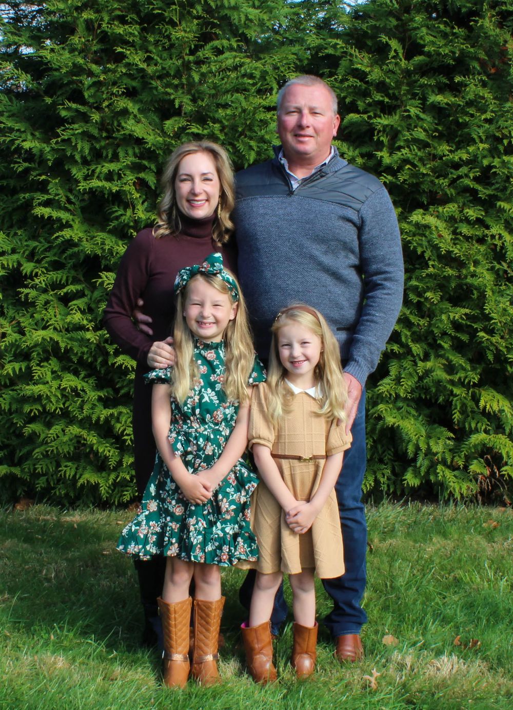 Family portrait taken outdoors against evergreen hedge with two young girls wearing boots in front.