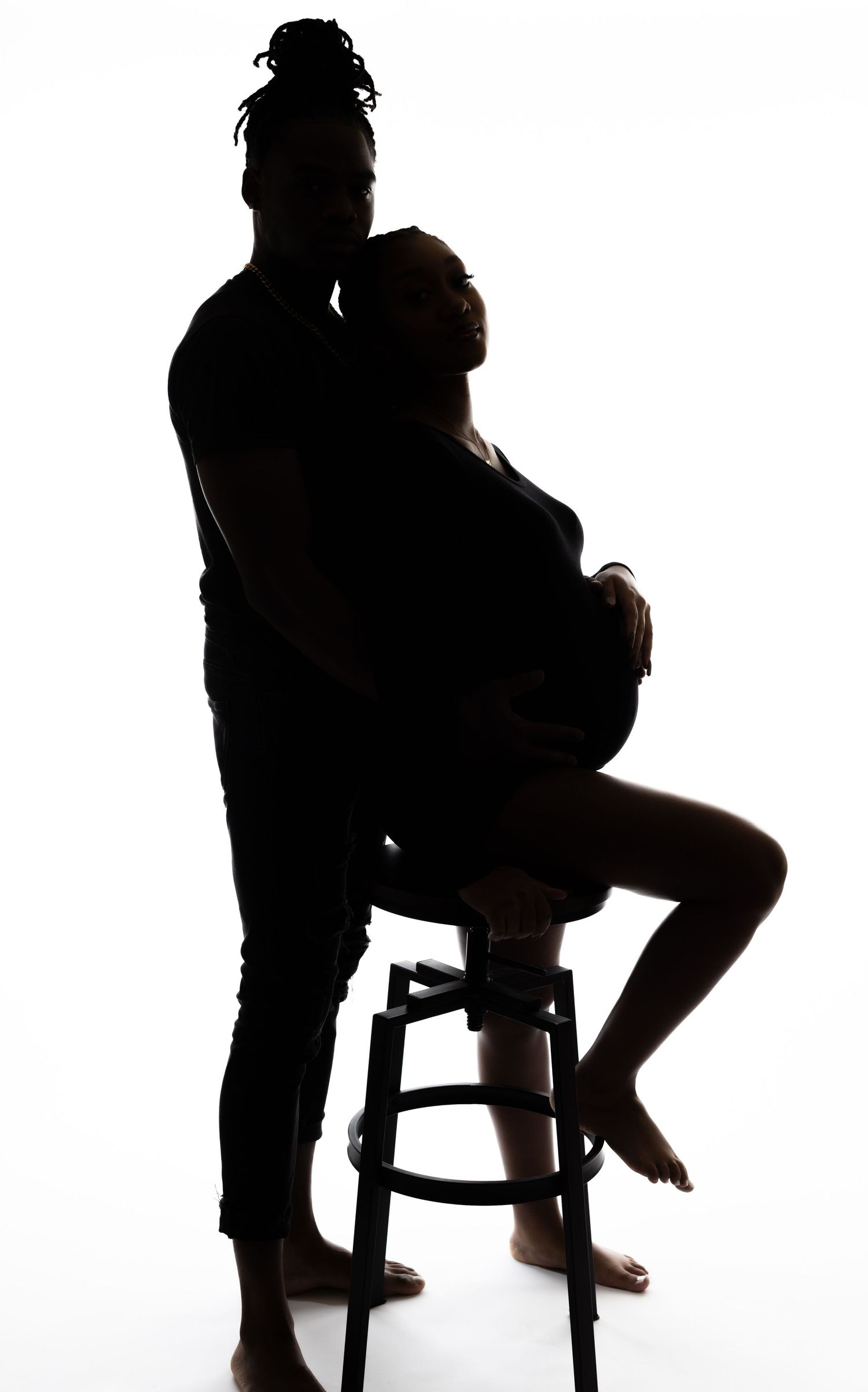 Artistic silhouette of couple during maternity shoot with woman seated on stool.