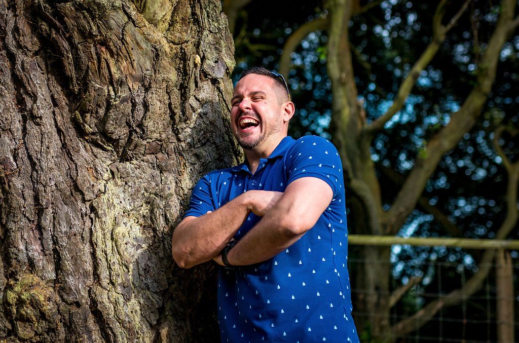 A candid portrait of someone laughing while leaning against a large tree trunk.