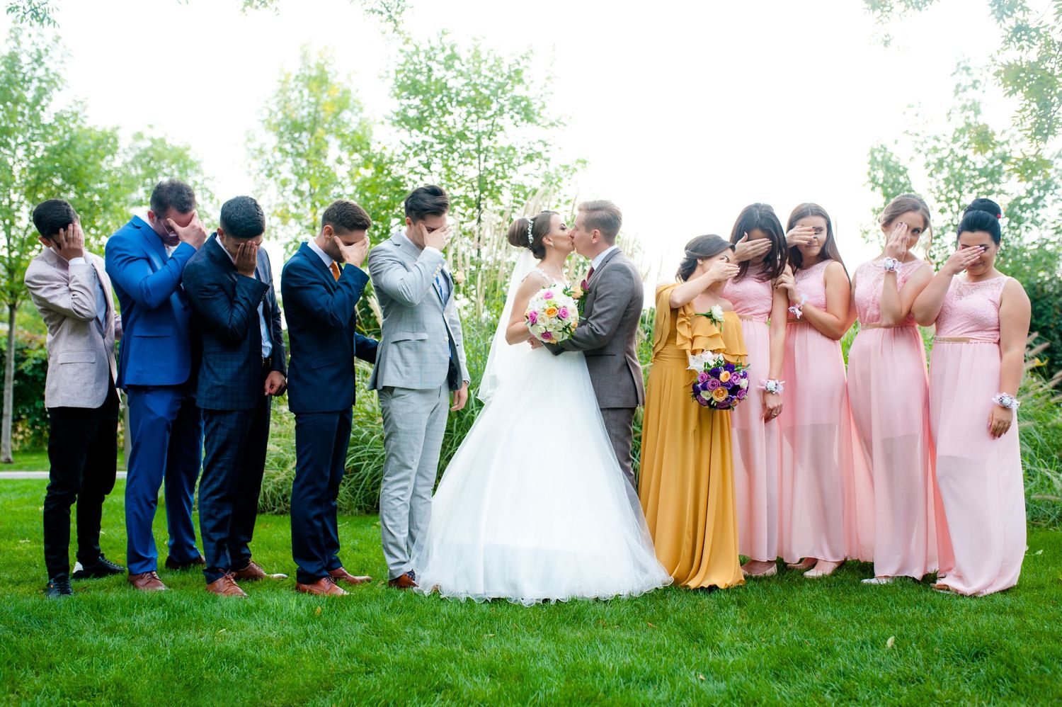 Wedding party in blue suits and pink dresses poses together on green lawn.
