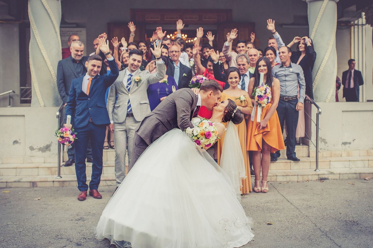 Wedding celebration with guests posing on steps while newlyweds share a romantic dip kiss.