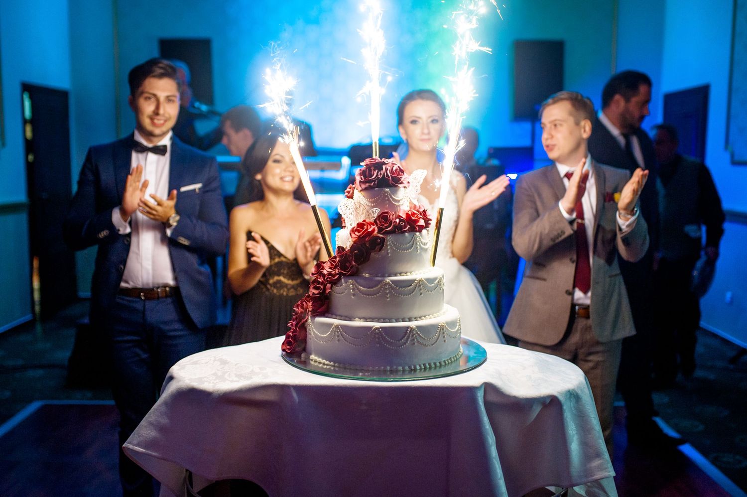 Wedding cake with sparklers glowing at reception while guests celebrate in blue-lit venue.