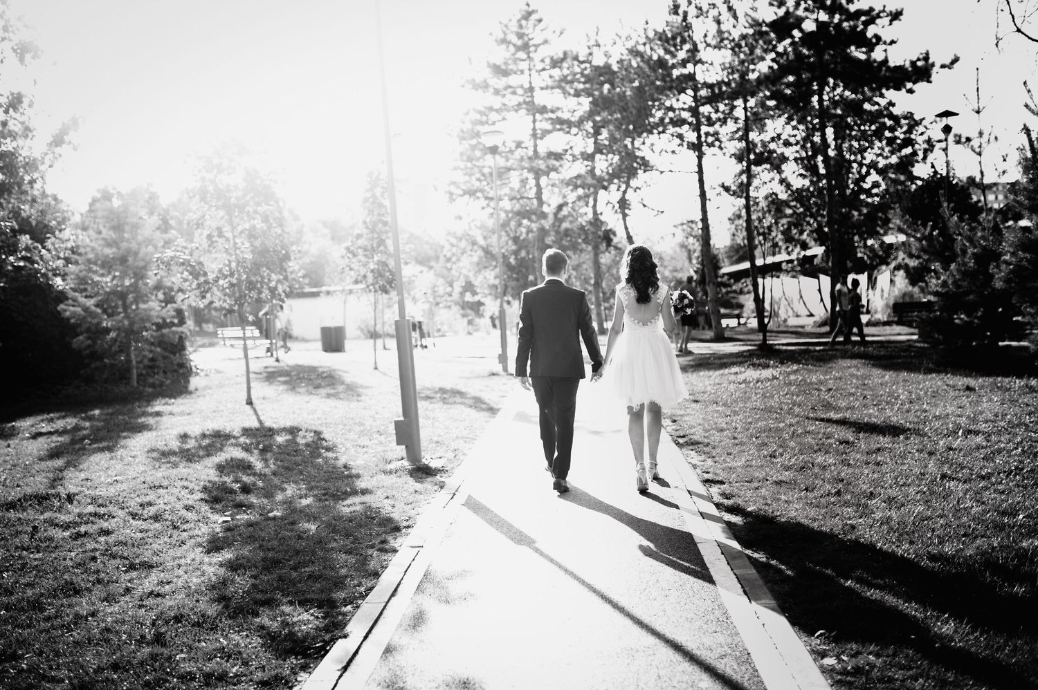 Black and white silhouette of newlyweds walking down sunny path among trees.