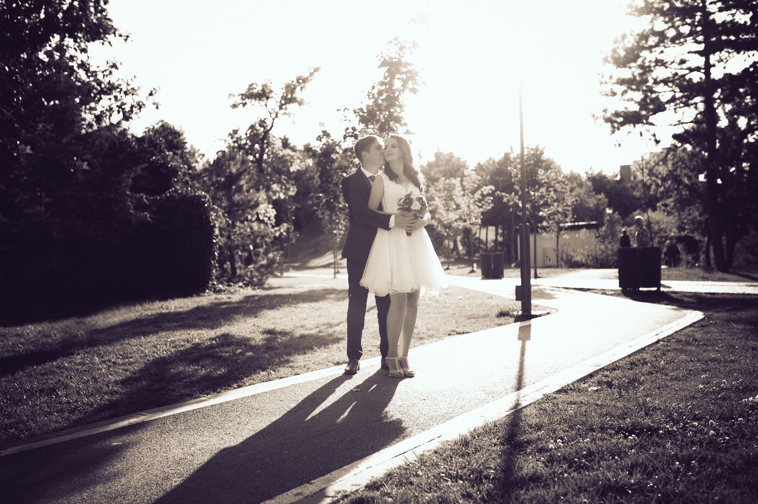 A black and white wedding photo of a couple walking on a sunlit path.