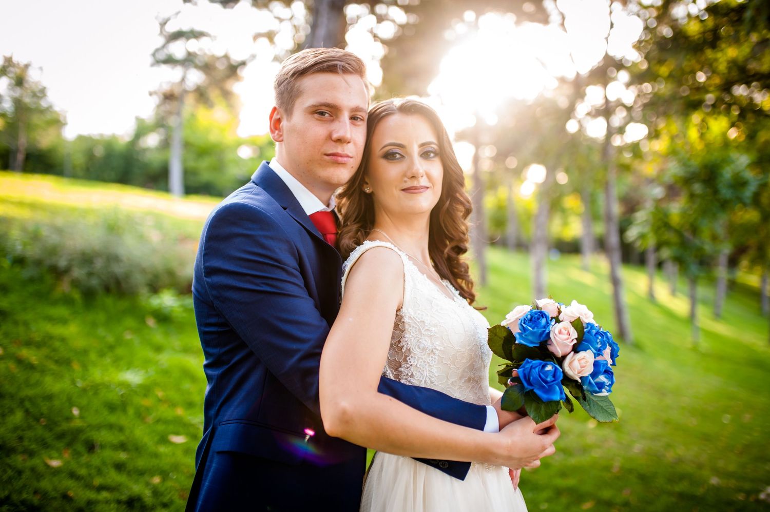 Newlyweds pose together in formal attire with blue bouquet during golden hour.