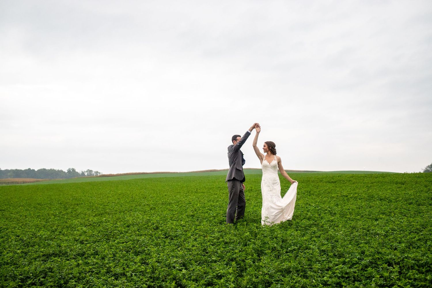 Bride and groom dancing joyfully in a lush green field under an overcast sky.