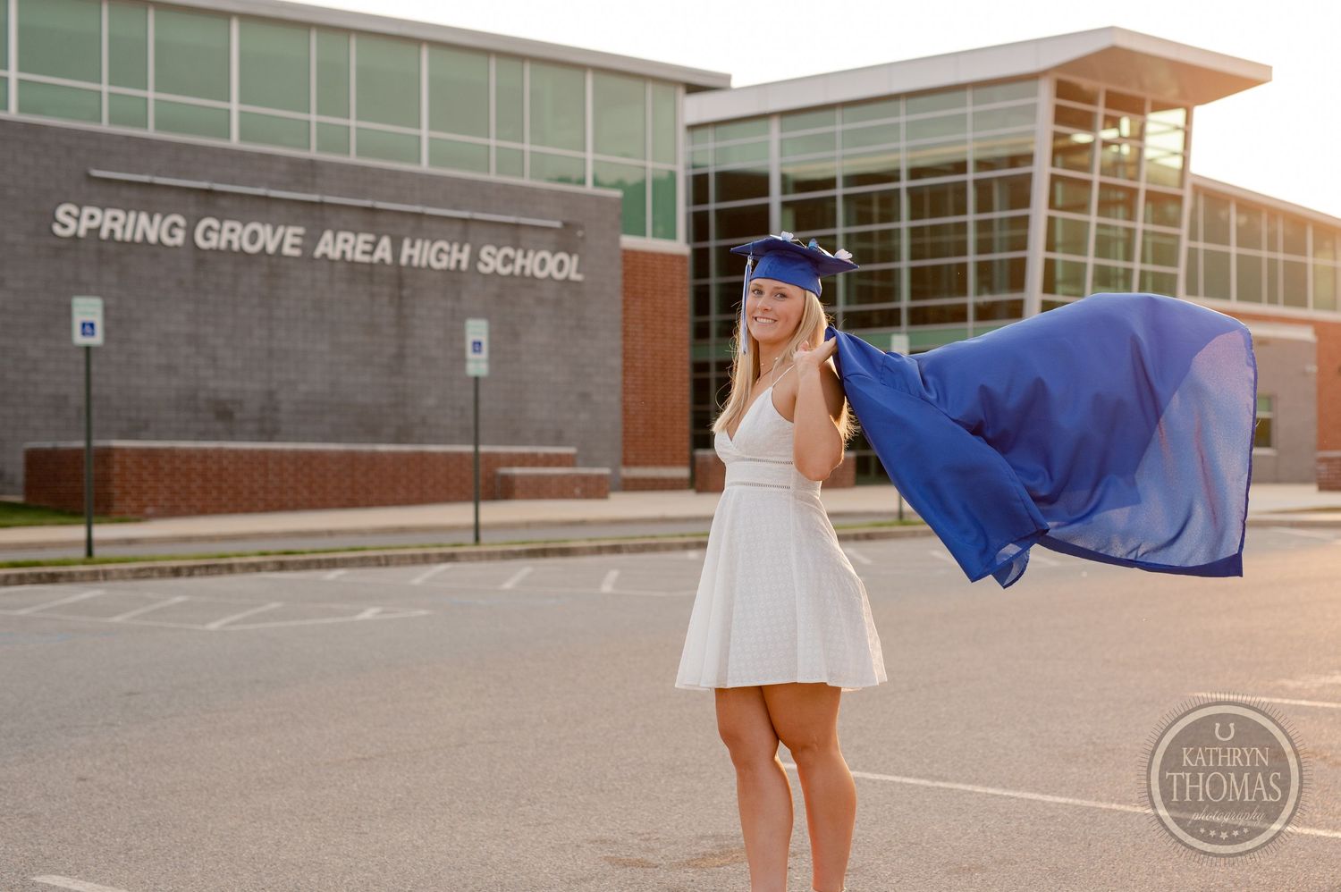 Graduate's blue gown flows in the wind as they pose outside their high school at sunset.