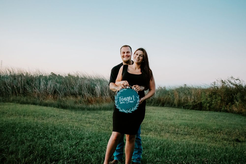 Expectant couple poses outdoors holding a teal announcement sign in a grassy field.