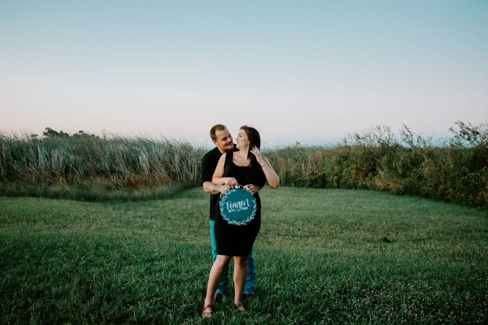Happy couple embraces while holding maternity announcement sign in sunset field setting.