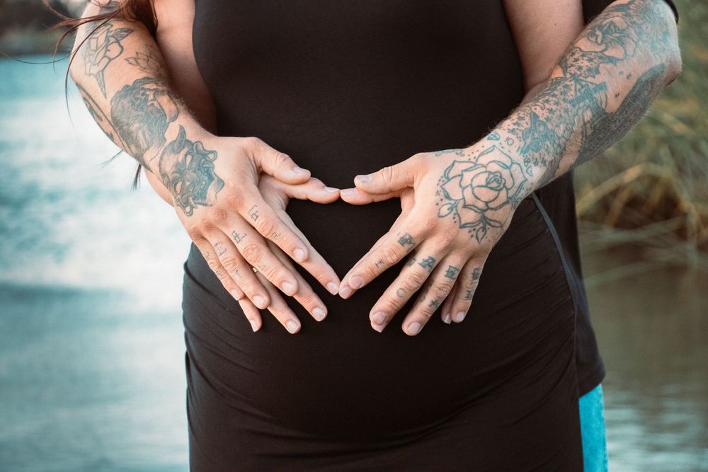 Tattooed hands form heart shape over pregnant belly in black maternity dress.