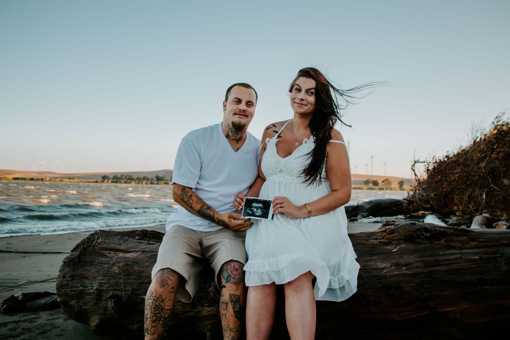 A romantic couple poses together on rocky shoreline at sunset with waves crashing in the background.