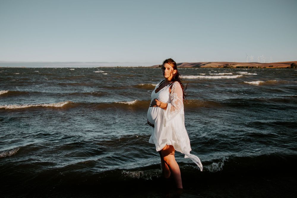Woman in flowing white dress stands on rocks by choppy ocean waters at sunset.