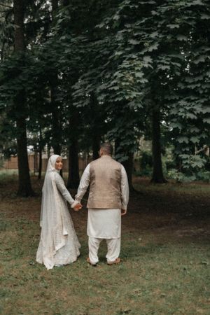 A couple in traditional wedding attire holds hands while walking through a forested park.