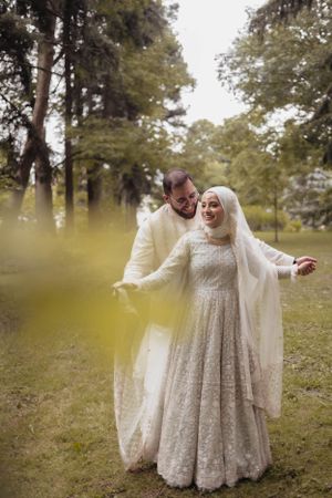 Newlyweds share a joyful moment in a forest setting wearing traditional white attire.