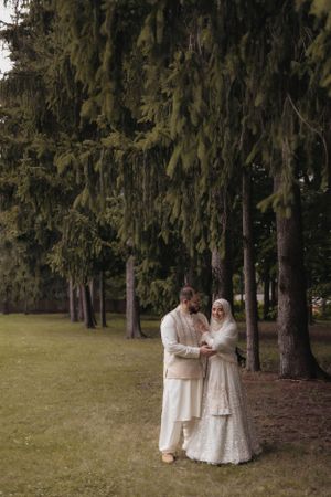 Couple in traditional white attire embrace among tall evergreen trees.