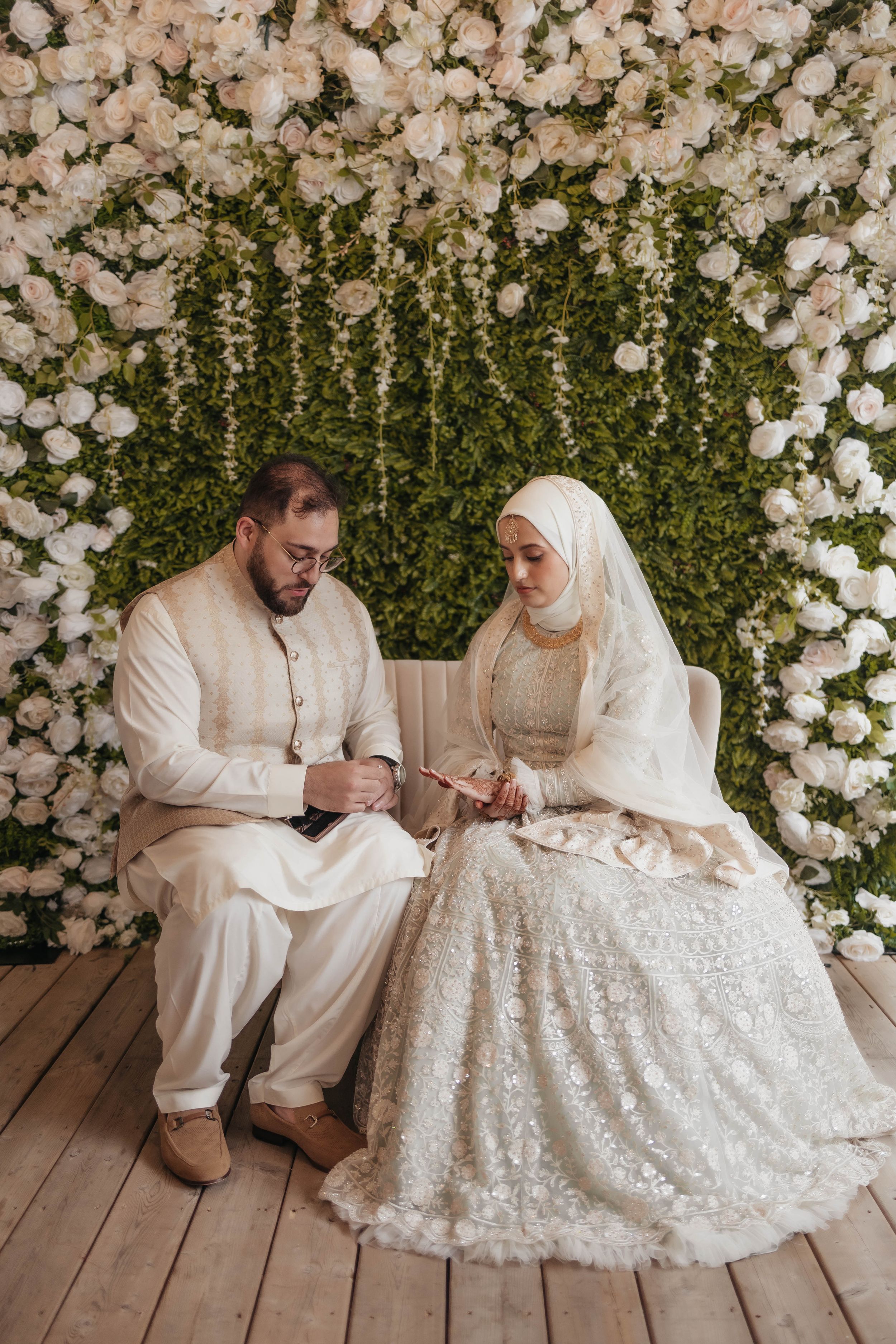 A wedding couple in traditional white attire seated beneath a floral arch with greenery.