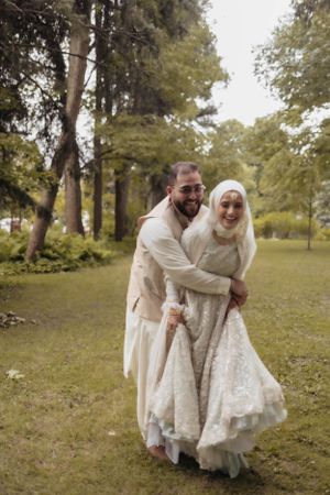 A couple in matching cream-colored traditional wedding attire share a joyful moment together in a garden setting.