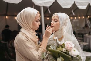 A tender moment during a wedding celebration as someone applies makeup to a bride holding a floral bouquet.