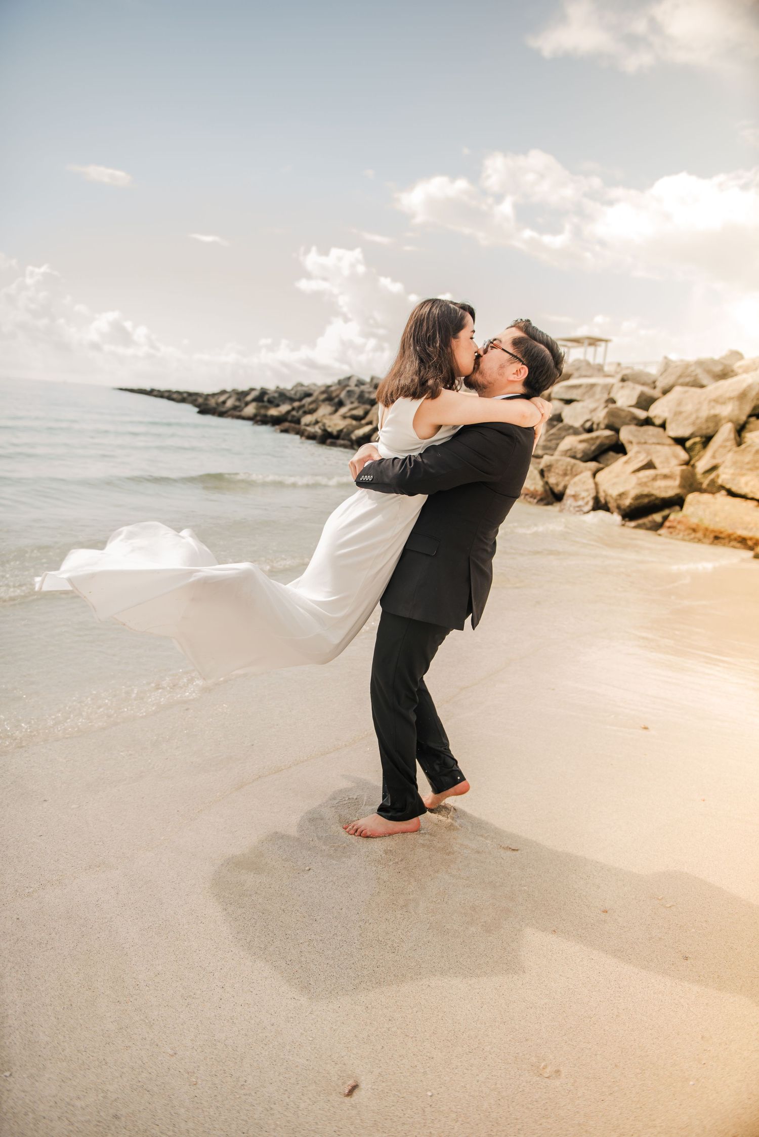 A couple embraces on a sandy beach with a rocky breakwater and cloudy sky in the background.