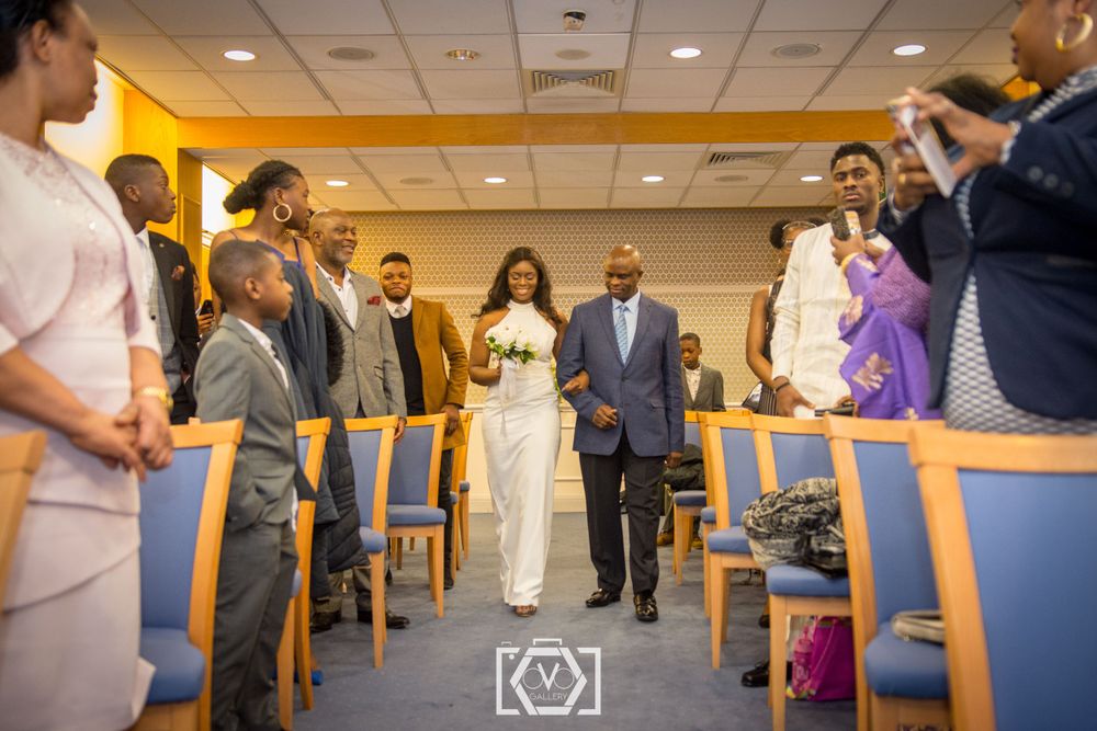 Bride walks down the aisle escorted by her father in a bright ceremony hall with guests watching.