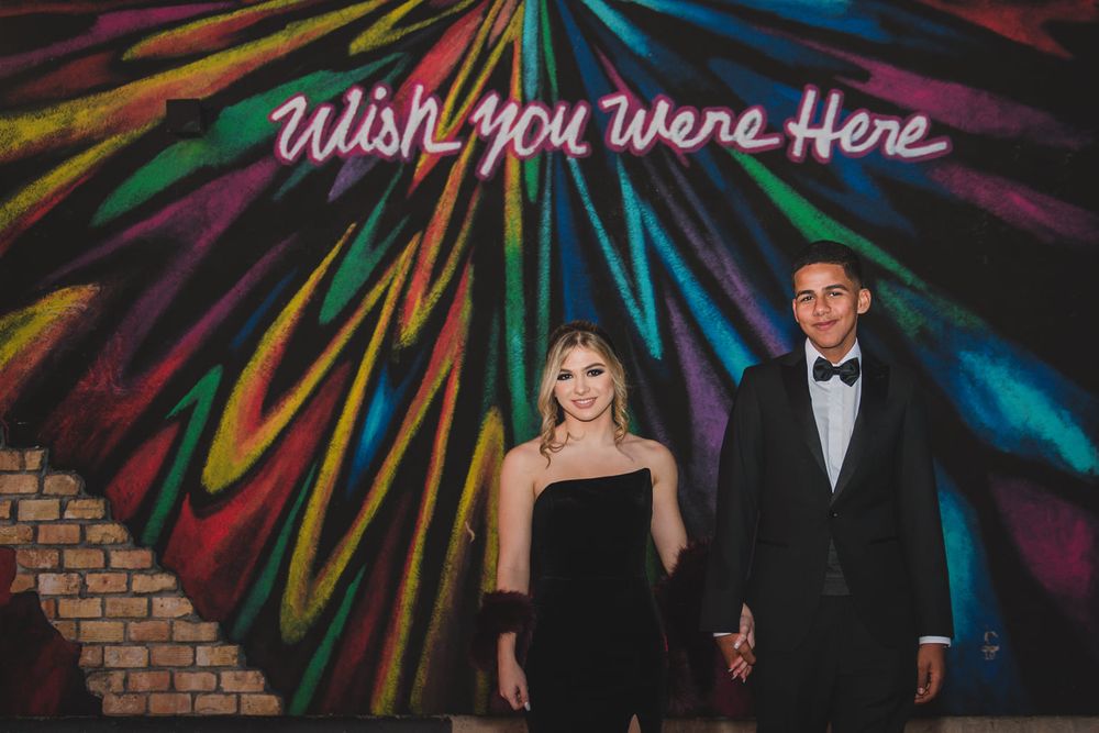 A couple poses beneath a neon 'Wish You Were Here' sign with colorful light streaks.