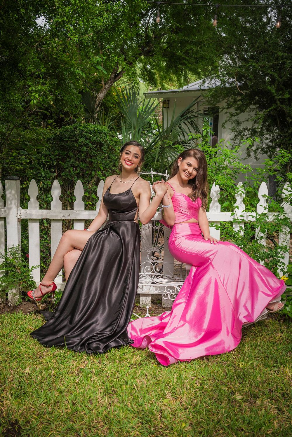 Two friends in formal dresses pose on white picket fence in garden, wearing black and pink gowns.