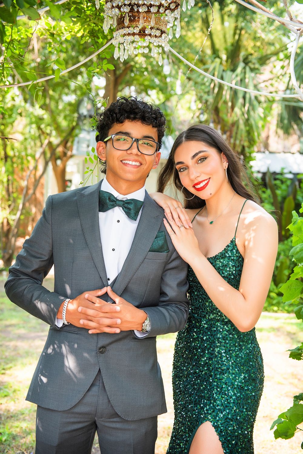 Formal couple pose under string lights in garden, wearing grey tuxedo and emerald sequin dress.