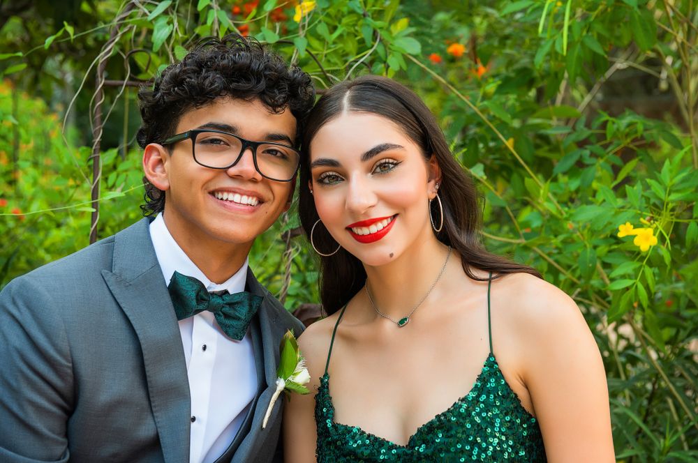 Formal portrait of prom couple in tuxedo and sparkling green dress against garden foliage.