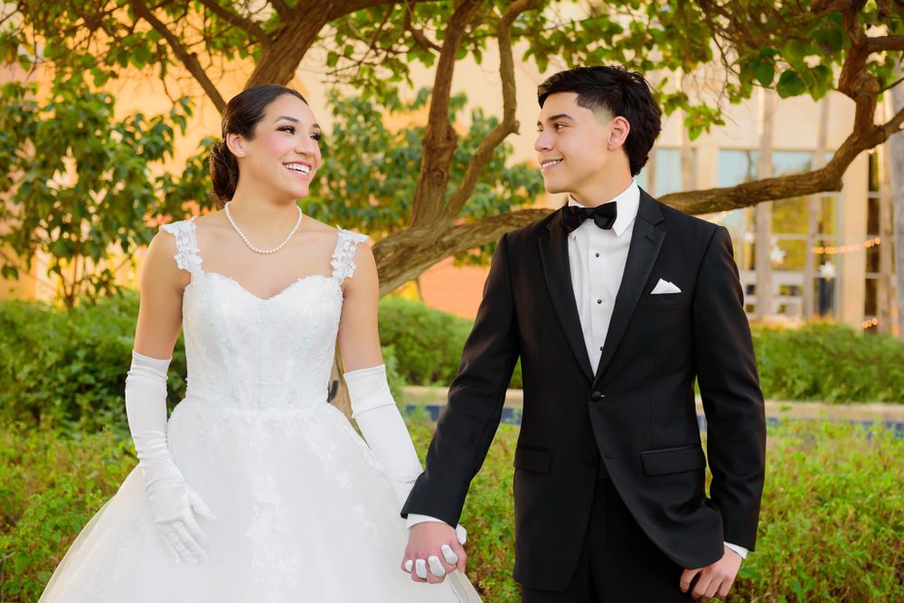 Wedding couple holding hands and smiling at each other under a leafy tree.