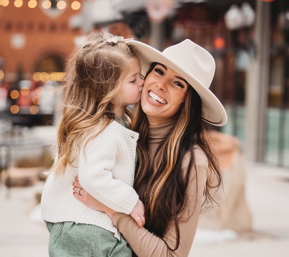 Candid outdoor portrait showing tender mother-daughter moment against urban backdrop with string lights.