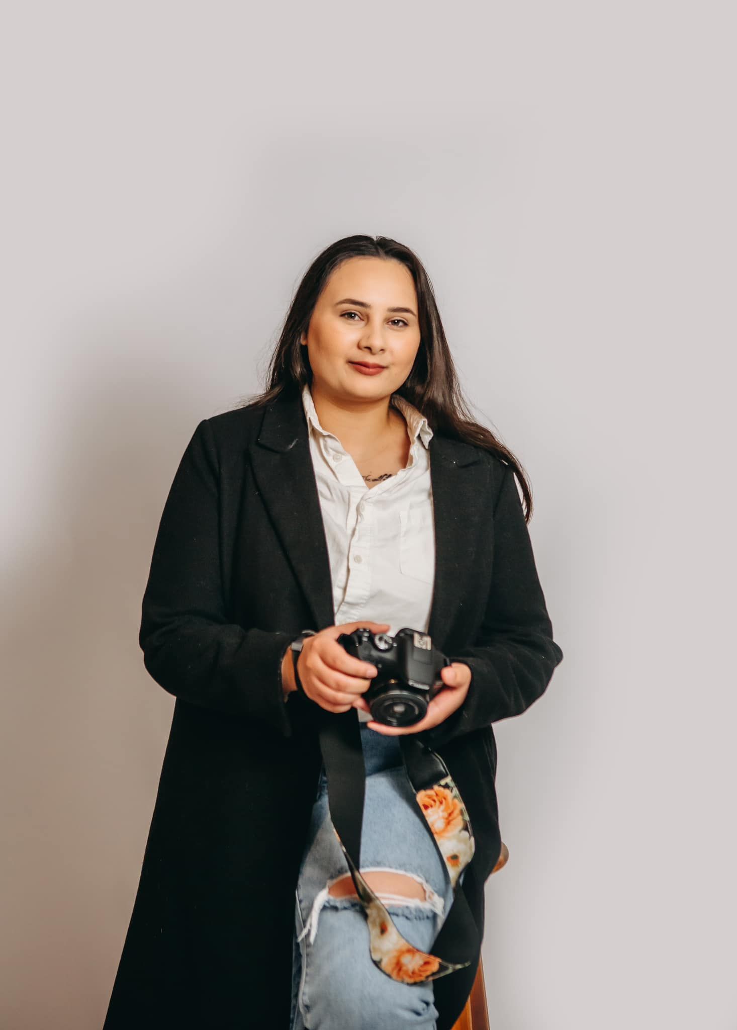 Professional photographer in black coat and distressed jeans holds camera against grey backdrop.