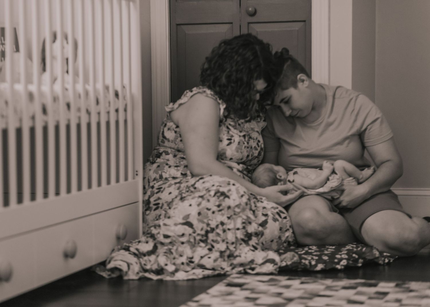 Two women sit on the floor with their daughter on a baby mat together in this black and white photograph with their newborn daughter.