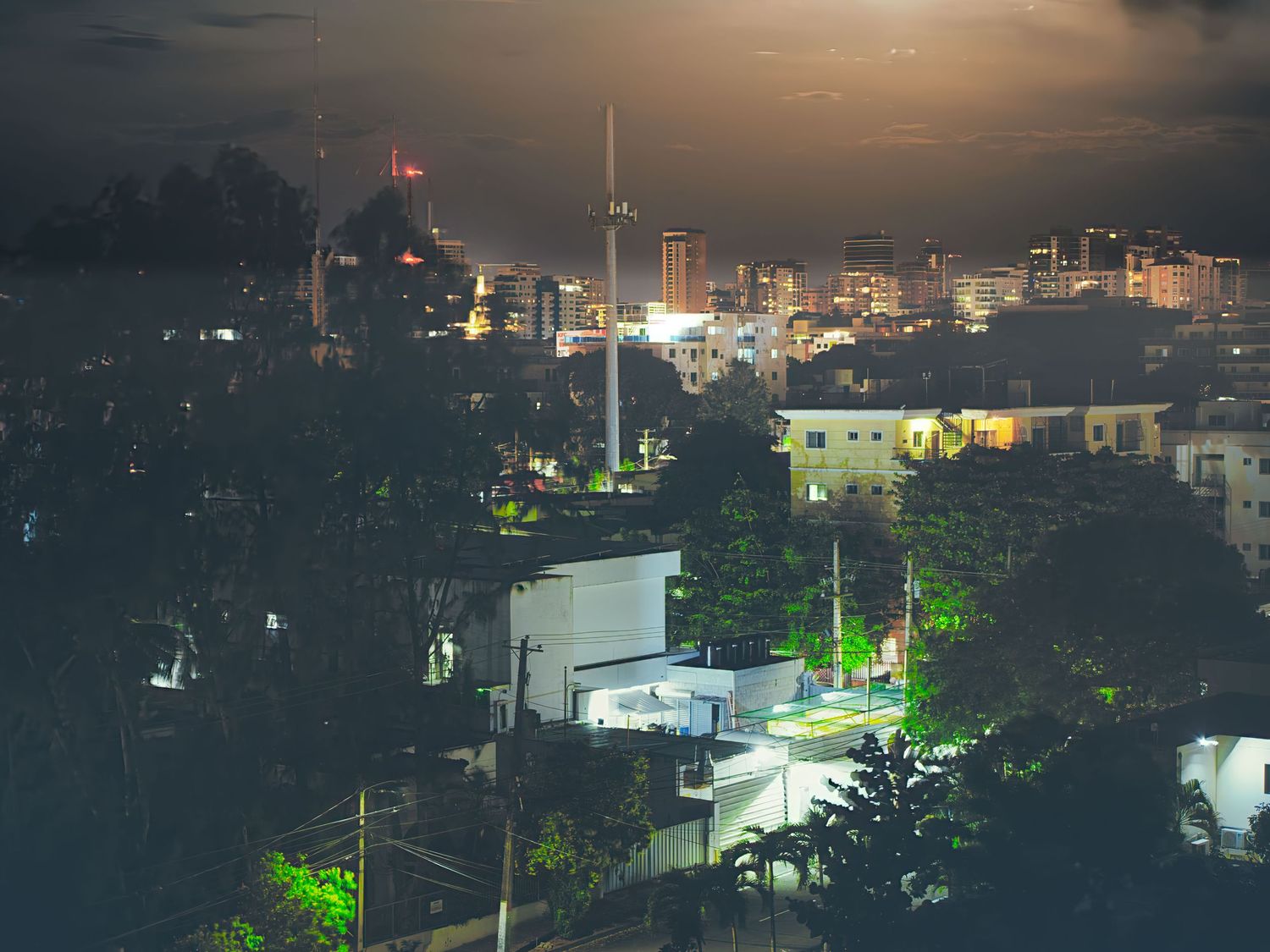 Aerial night view of a city skyline with glowing lights and tall buildings under cloudy sky.