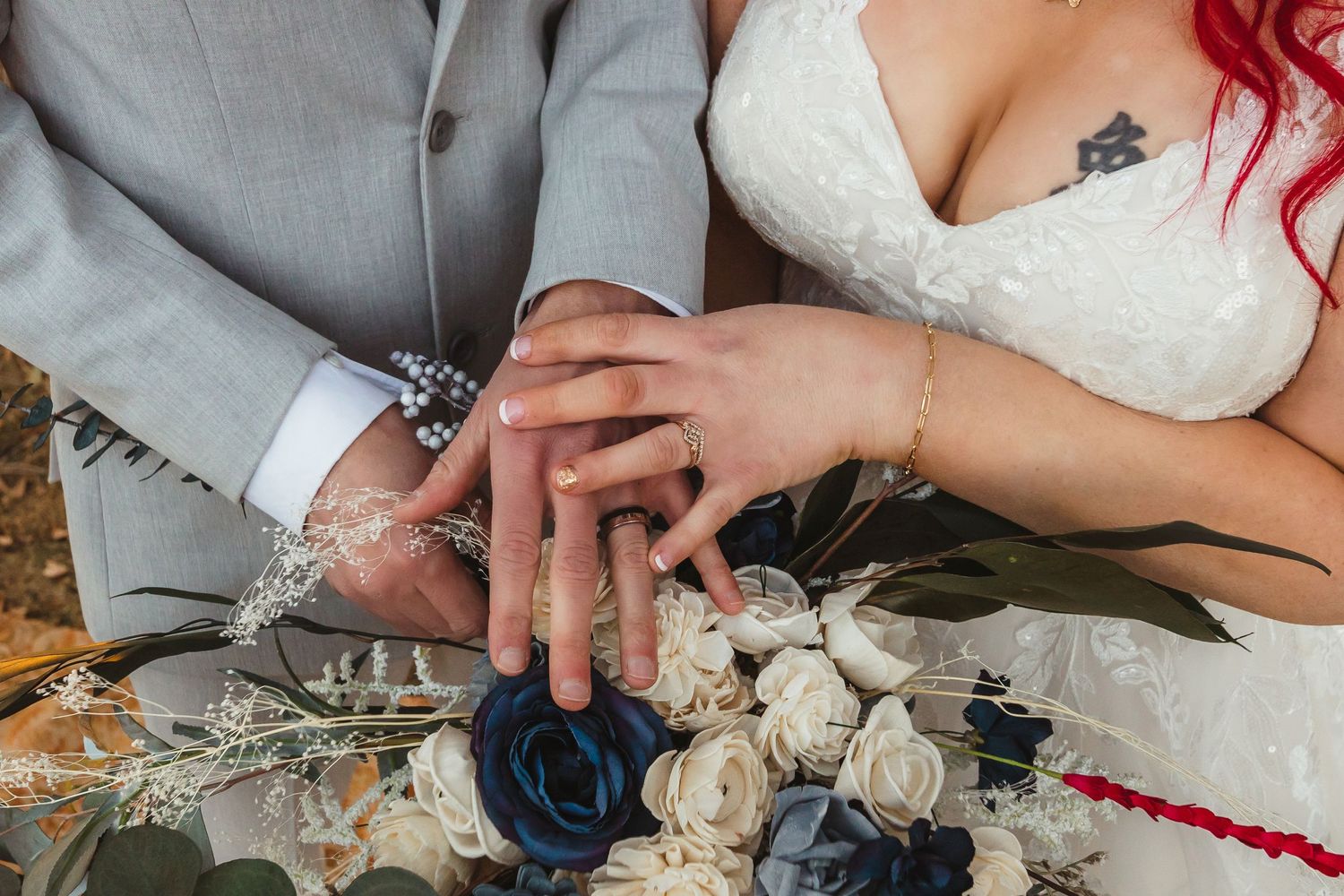 Close up photo of wedding rings with hands resting on a bouquet of navy blue and white roses.