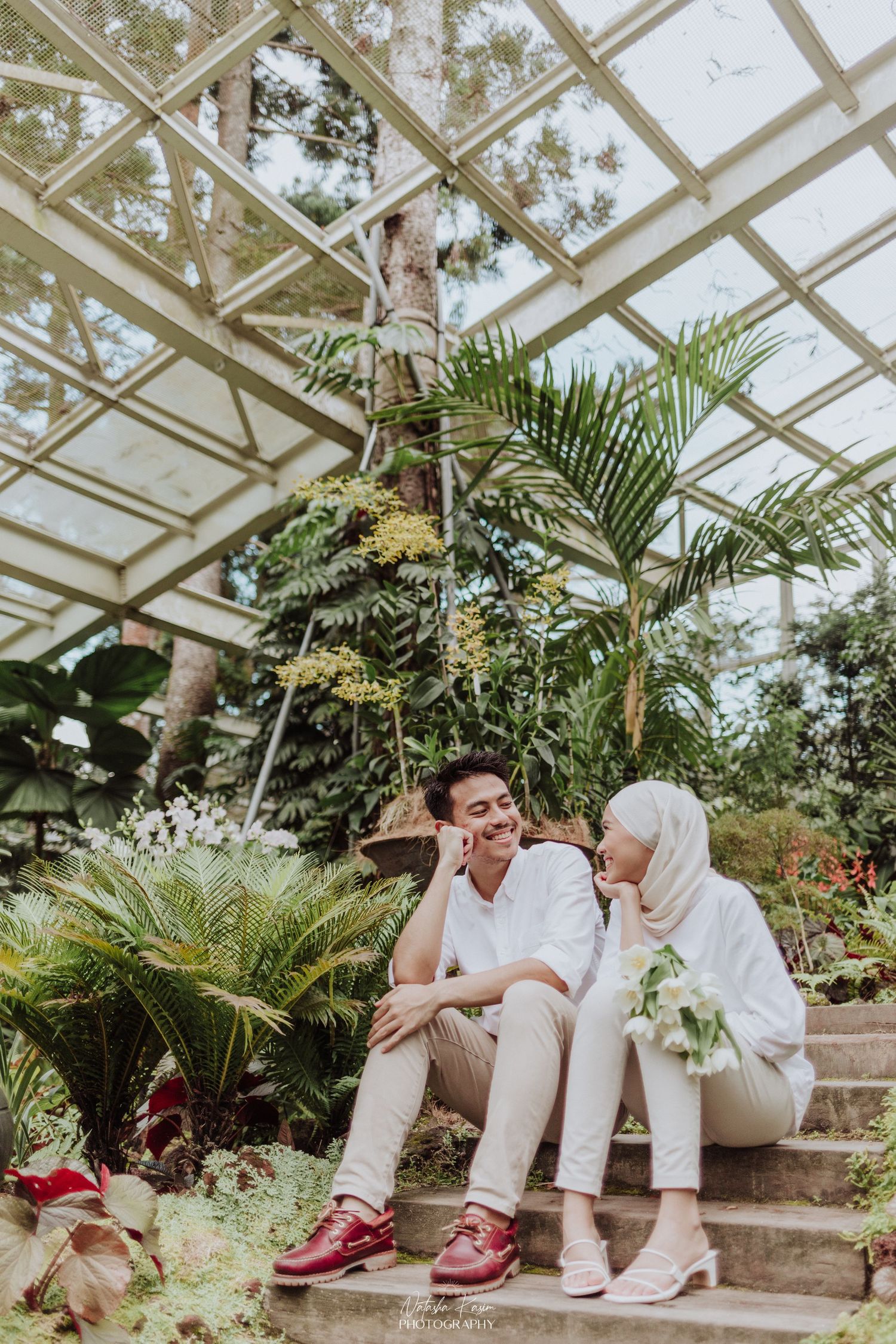 A couple sits on greenhouse steps surrounded by tropical plants and glass ceiling architecture.
