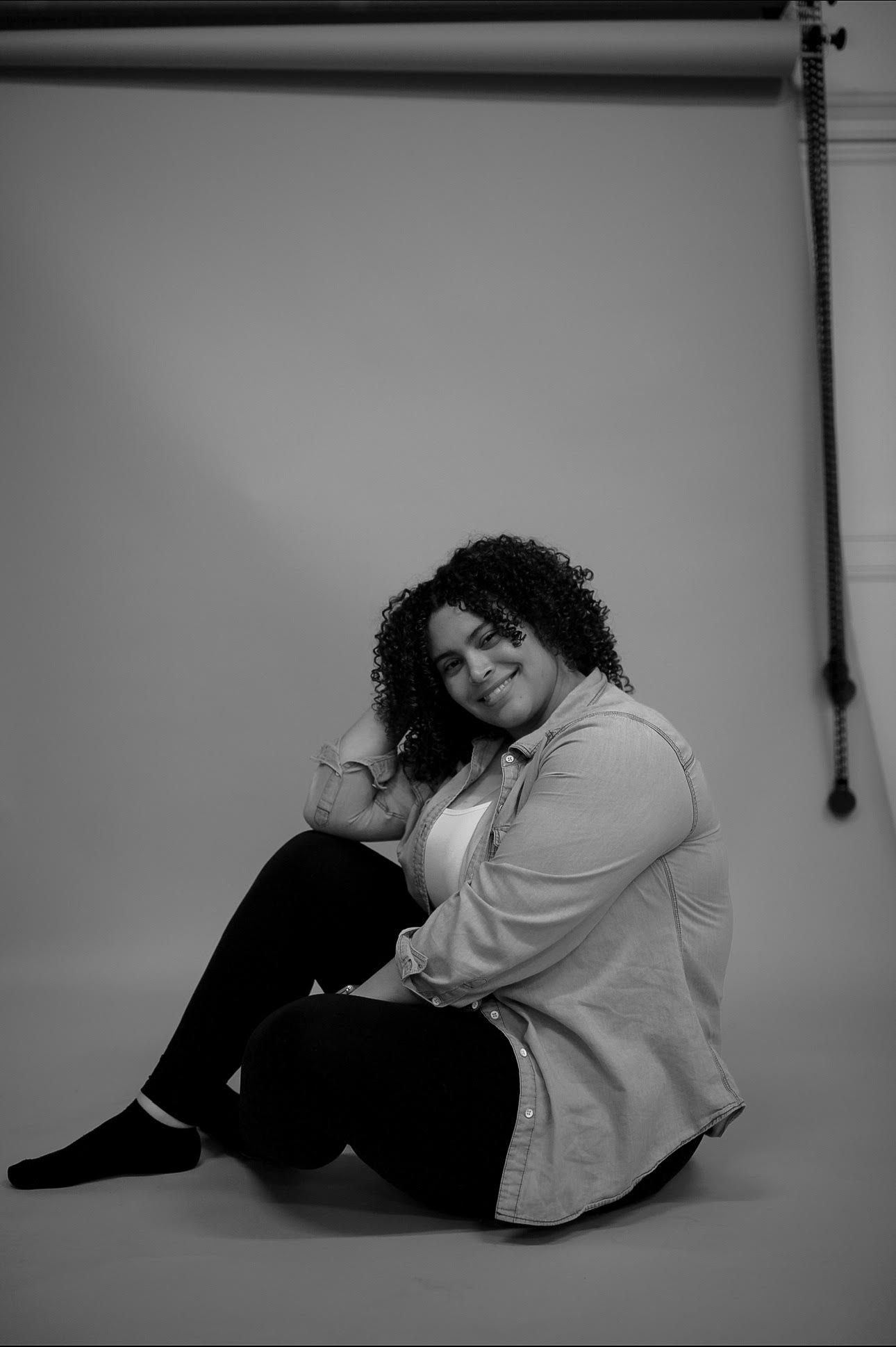 Black and white portrait of person in casual attire sitting against studio backdrop.