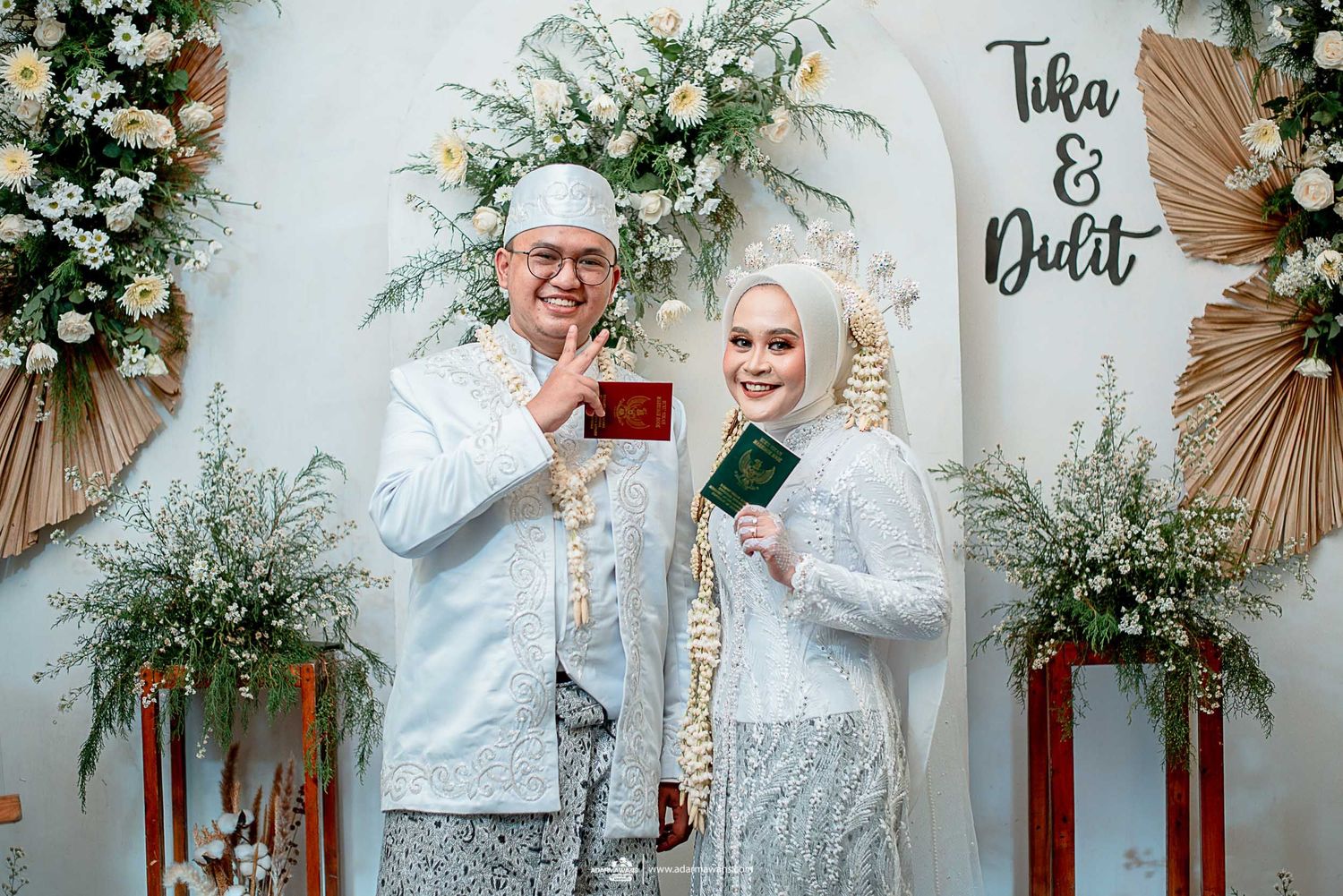 Wedding couple in traditional white attire posing with documents against floral arch backdrop.