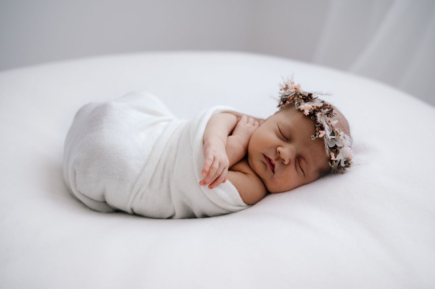 Peaceful sleeping newborn swaddled in white fabric wearing a delicate floral crown on soft surface.