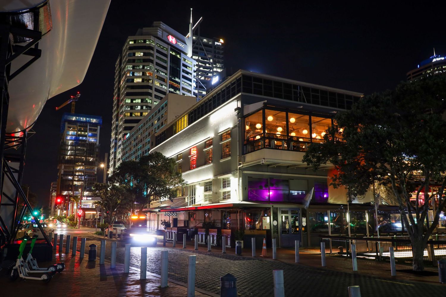 A vibrant city street scene at night, with high-rise buildings, lit-up windows, and colourful lights reflecting off the wet pavement.